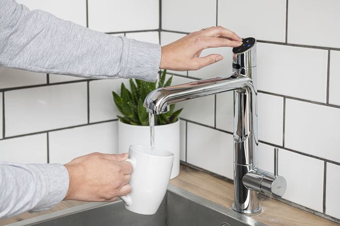  A man pouring boiling water into a mug from a kitchen tap, next to a plant.